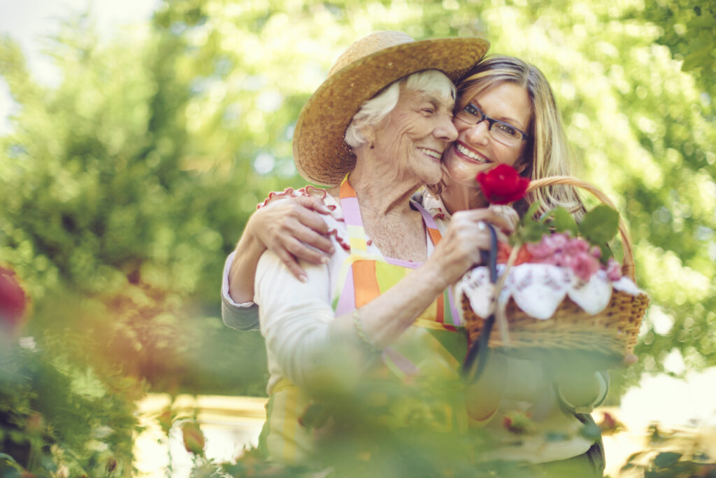 senior mother and adult daughter outside smiling in rose garden summer sunshine